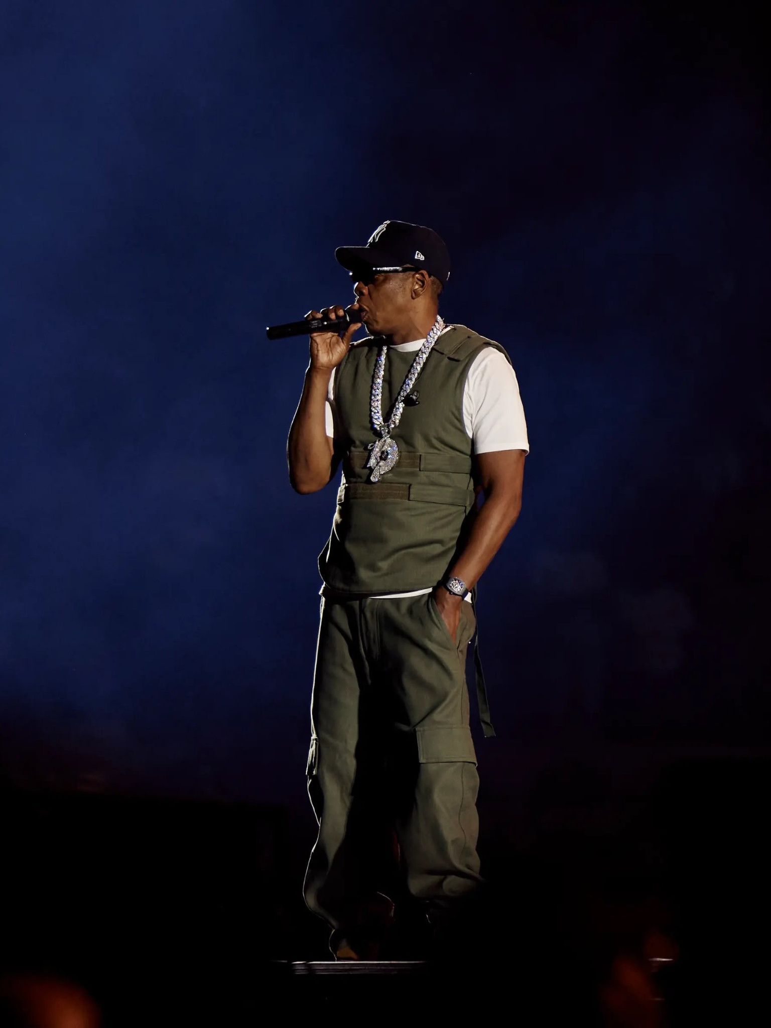 Jay-Z performs in an olive vest and white T-shirt, wearing a broad stone-set necklace and Roc-A-Fella pendant.