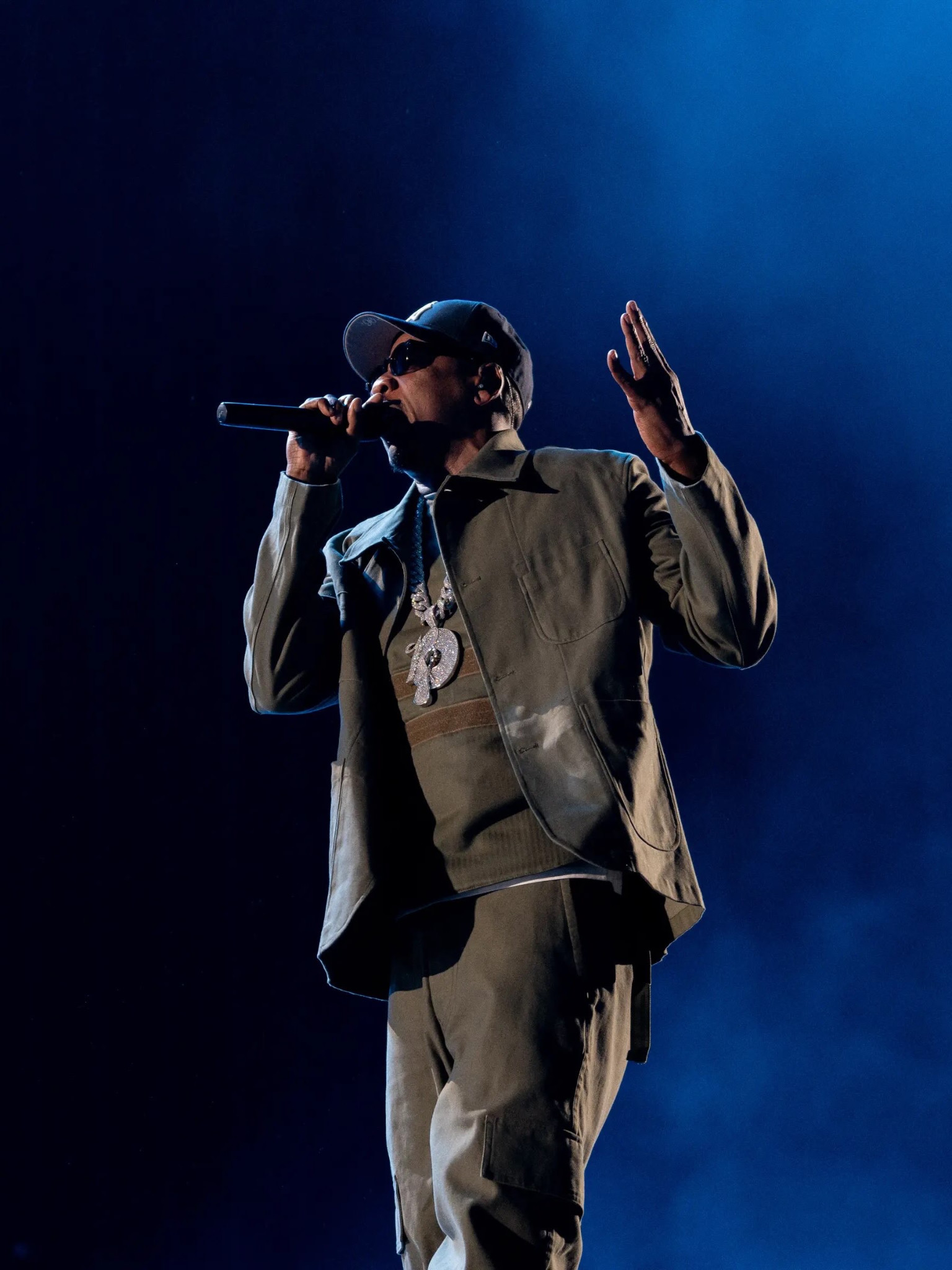 Jay-Z performs with a microphone and one hand raised, wearing a Roc-A-Fella pendant with an olive jacket.